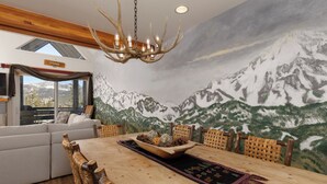 Un comedor con una mesa de madera, sillas tejidas y un mural de montañas en la pared.