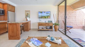 Una cocina moderna con armarios de madera, microondas integrado y una isla grande. Hay un comedor con mesa y sillas de madera, una planta en maceta y un cuadro con una escena de playa.