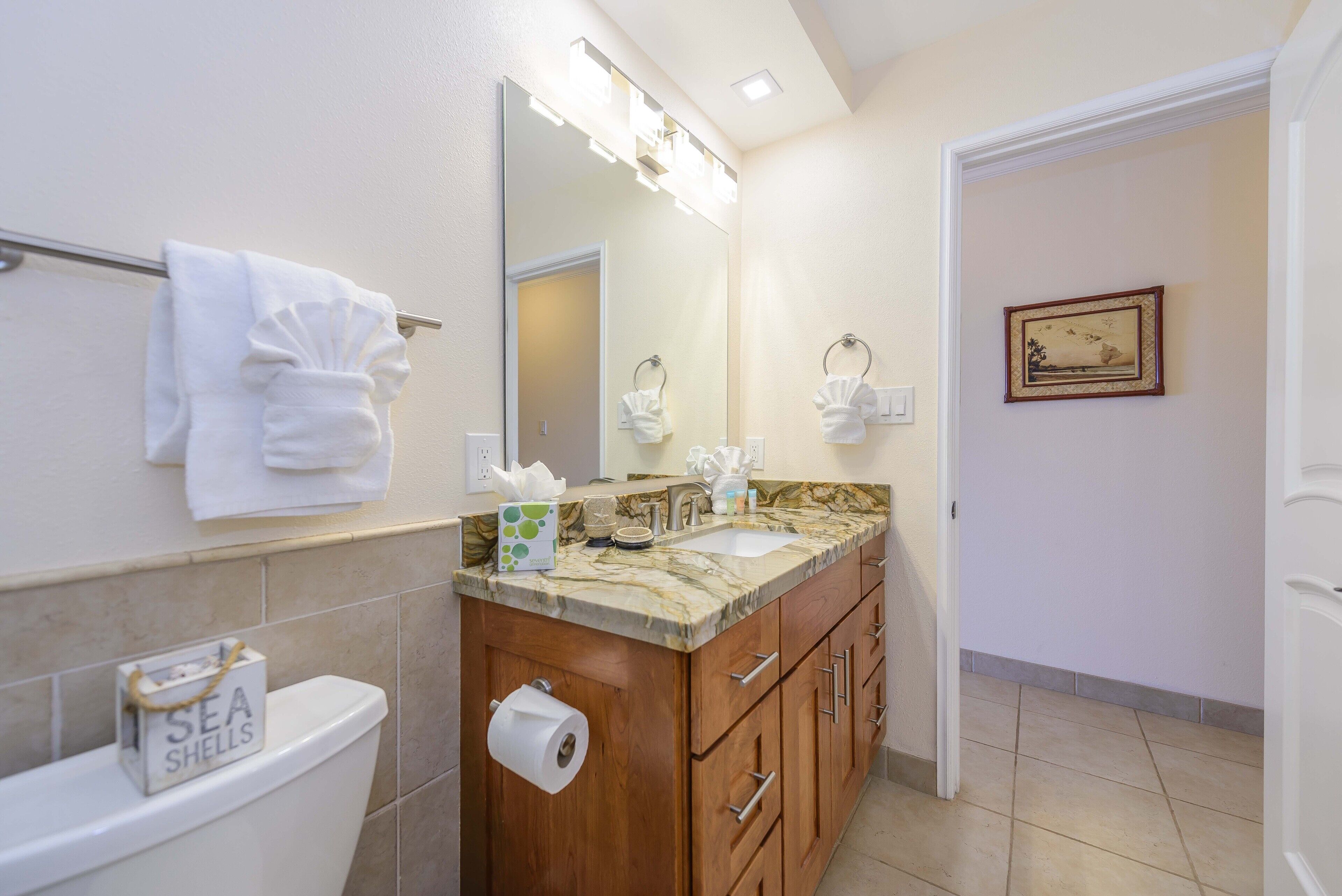 A bathroom with a marble countertop, wooden cabinets, a large mirror, and a toilet with a 'SEA SHELLS' sign.