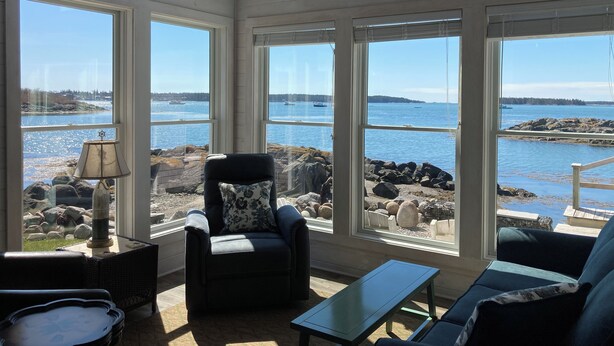 A sunlit room with large windows overlooking a rocky shoreline and calm water. - Beautifully Renovated Ocean Front Cottage With Spectacular Views.. (Jonesport)