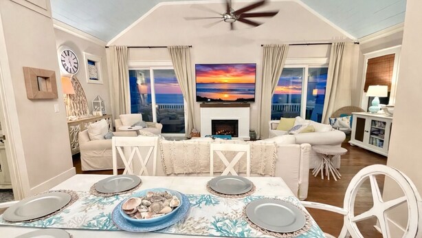 Een ruime woonkamer met een open haard, een eethoek met een tafel voor vier personen, en een groot raam met uitzicht op een zonsondergang.