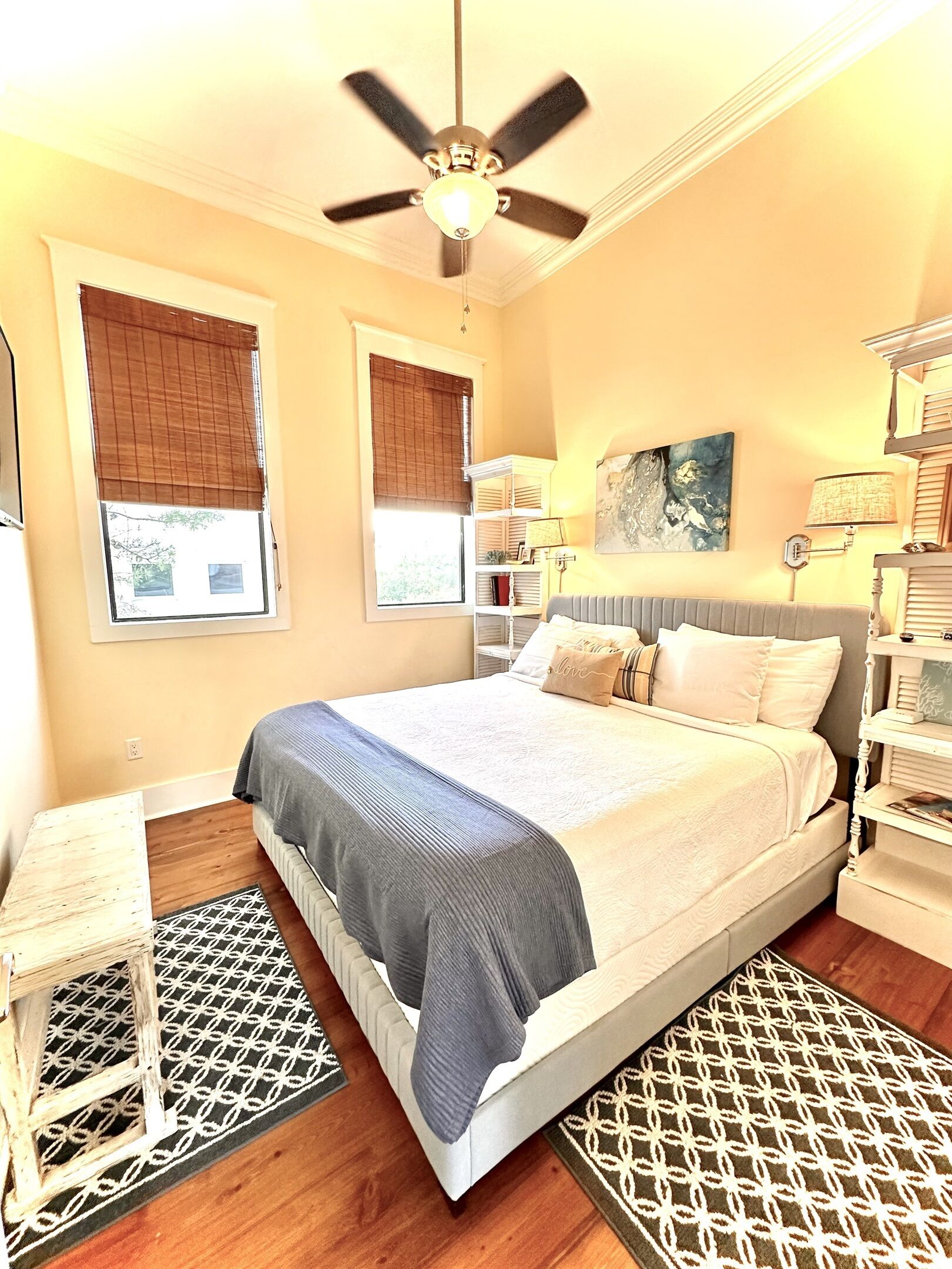A bedroom with a bed, a ceiling fan, two windows with blinds, a bedside table, a lamp, and a painting on the wall.