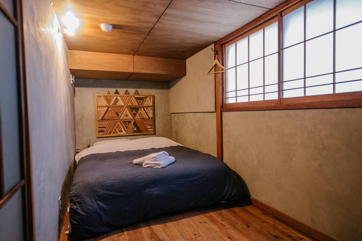 A bedroom with a wooden ceiling, a bed with a dark blue bedspread, a wooden headboard with a geometric pattern, and a window with wooden frames.