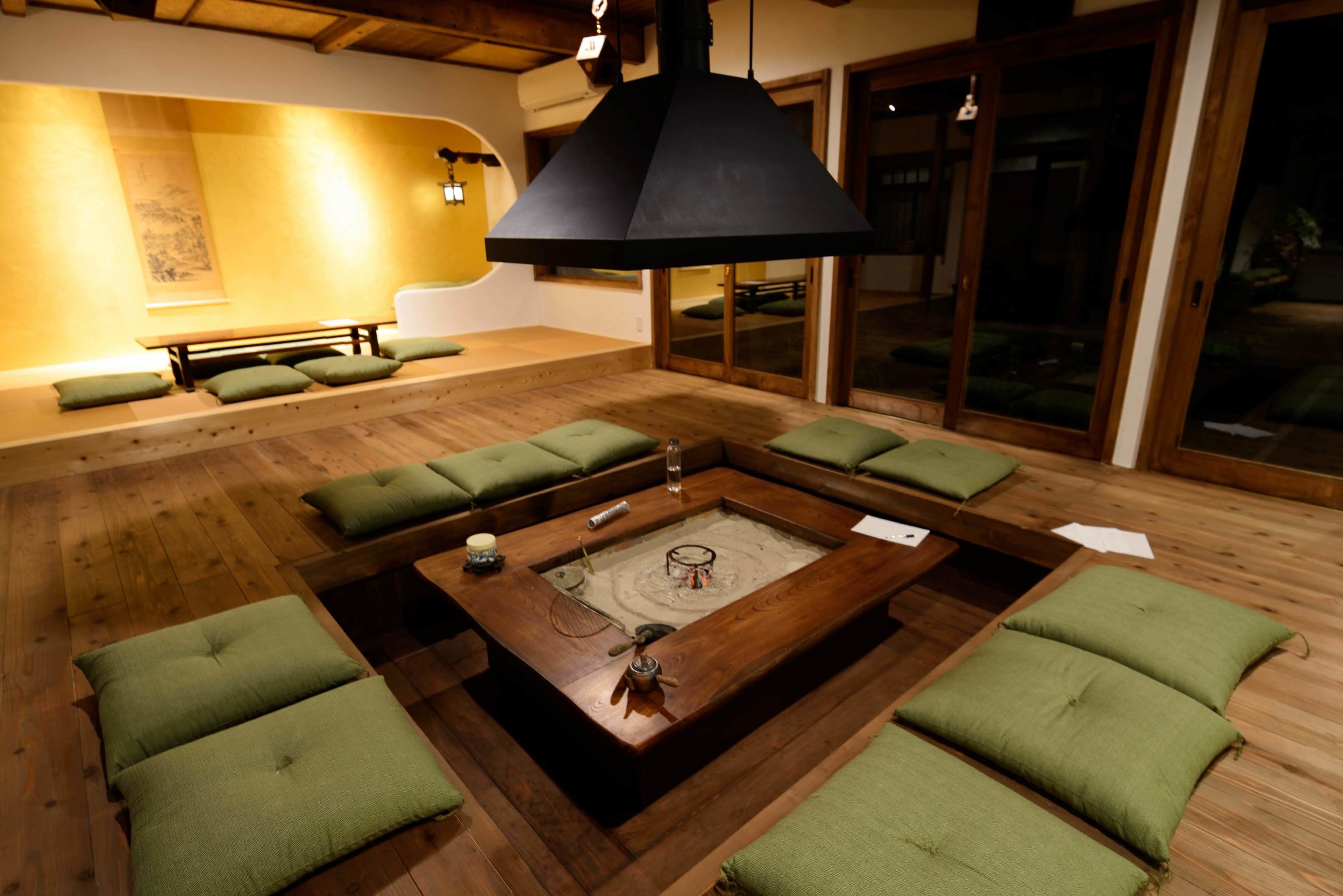 A room with a central wooden coffee table, green cushioned seating, and a large black pendant light.