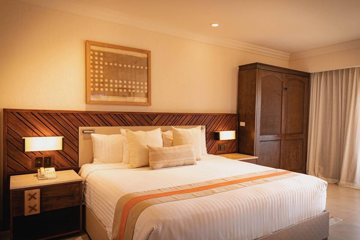 A hotel room with a large bed, wooden headboard, two bedside tables with lamps, a wardrobe, and a framed artwork on the wall.