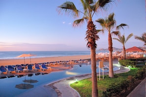 A poolside area with lounge chairs and umbrellas, a beach in the background, and palm trees.