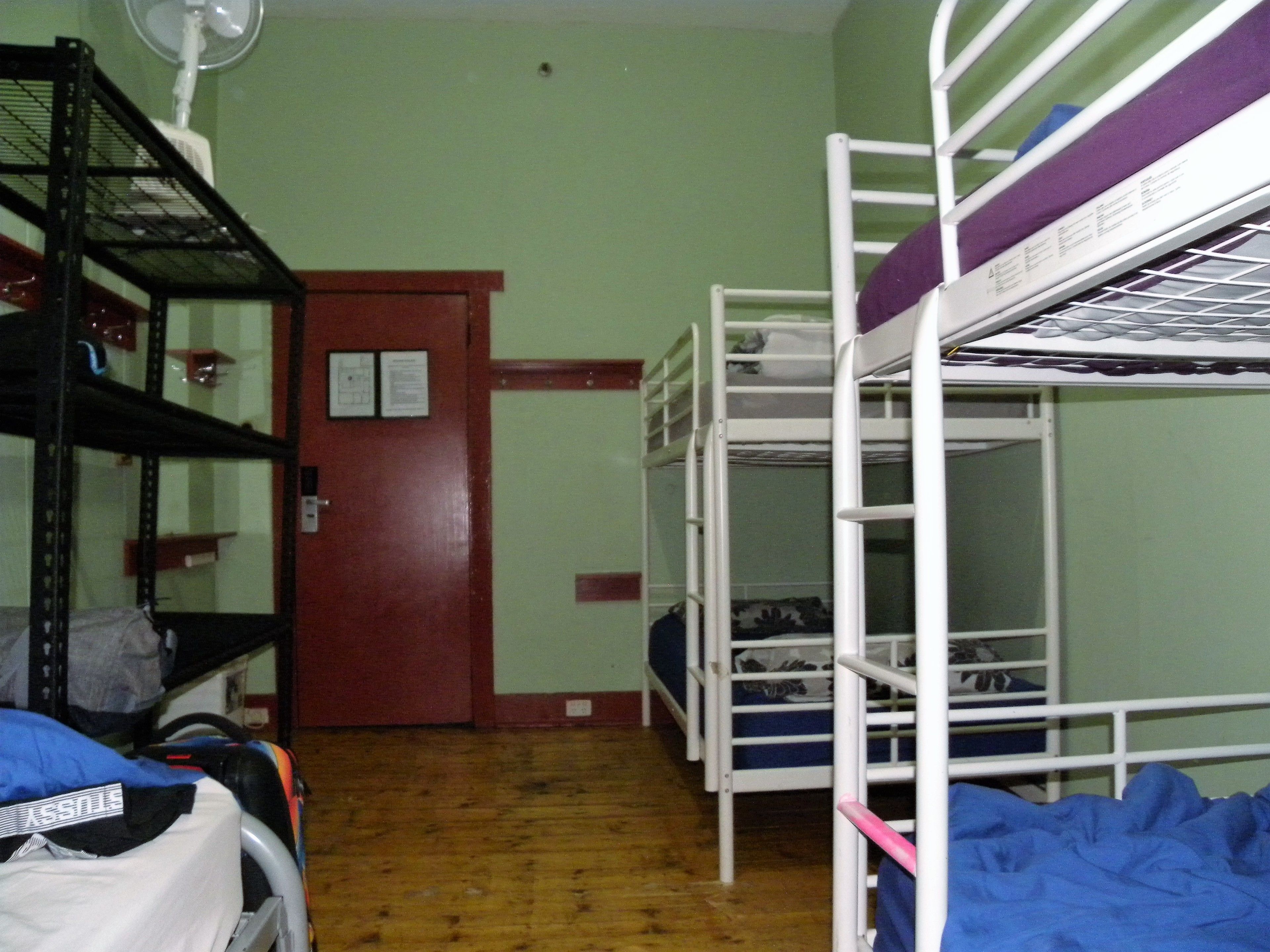 A dormitory room with bunk beds, a wooden floor, a door, and a fan on the wall.