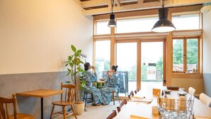 Un restaurante con mesas y sillas de madera, ventanas amplias y una planta.
