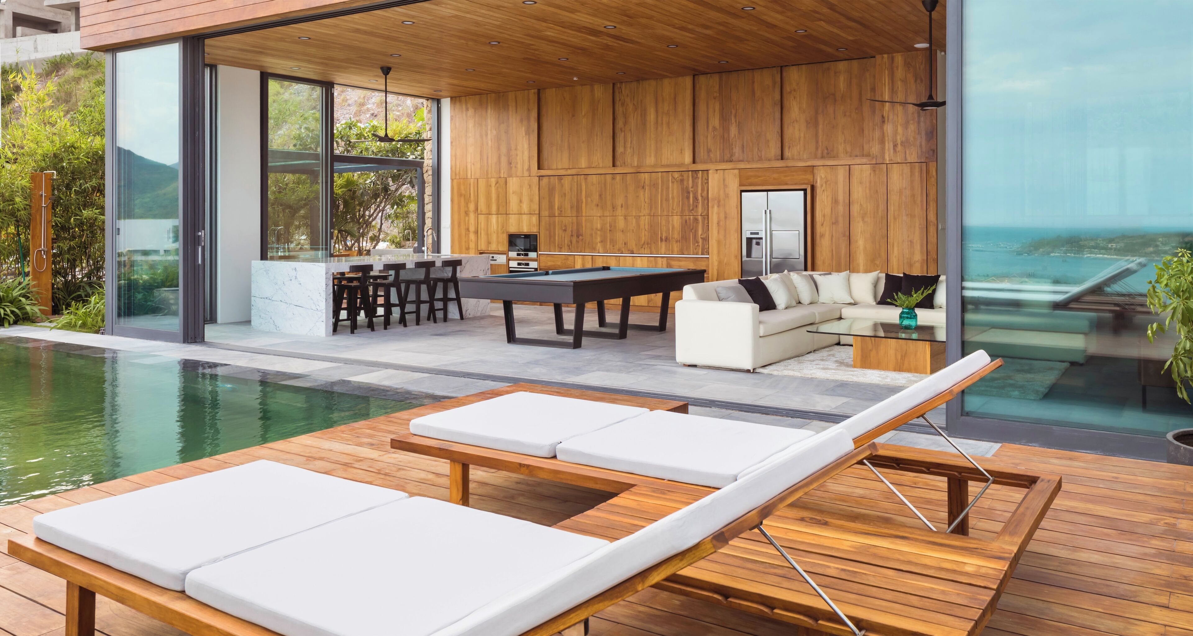 Ein moderner Außenbereich mit Pool, Holzterrasse, Liegestühlen, einem Billardtisch und einer Sitzgruppe mit Meerblick.