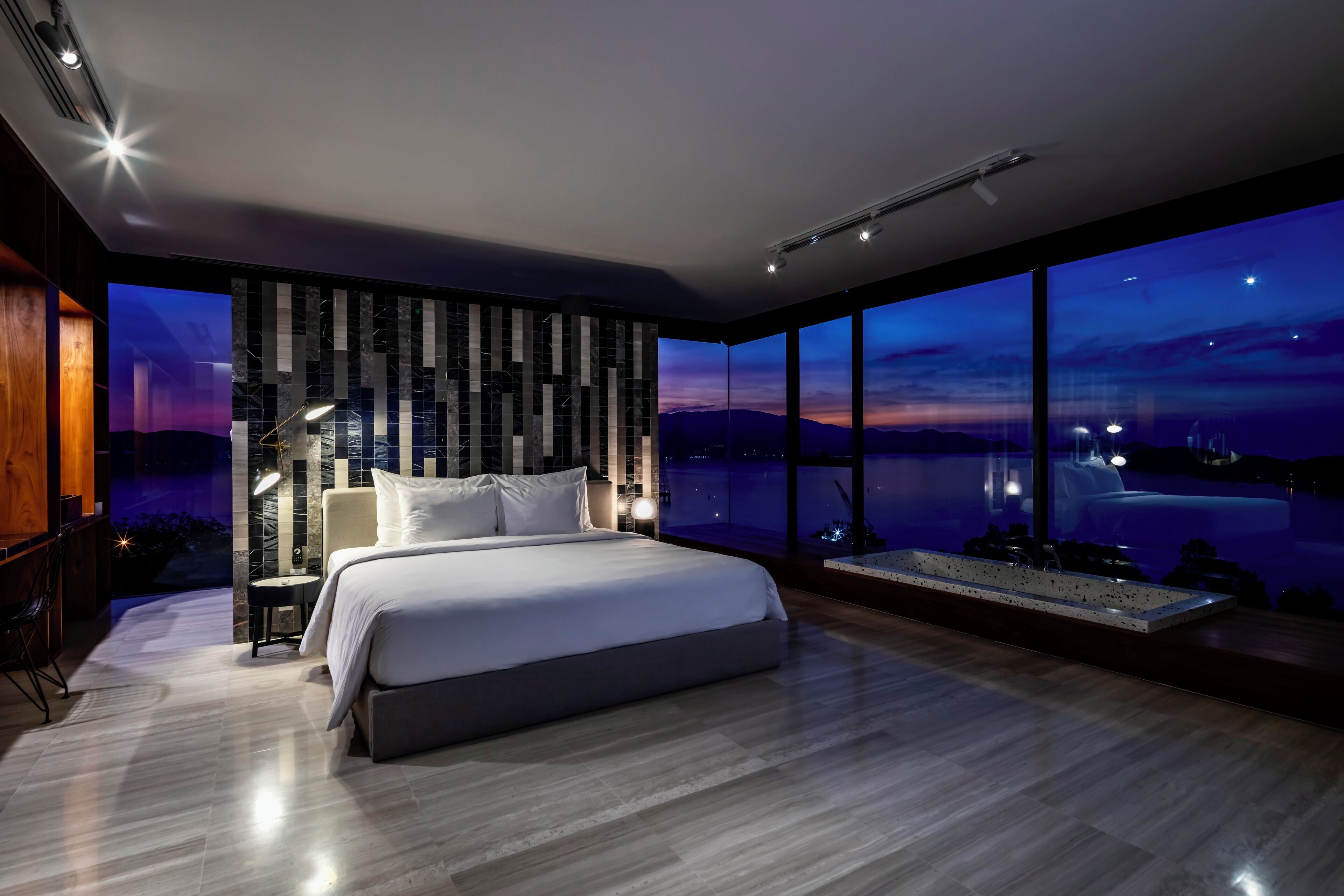 Un dormitorio moderno con una cama grande, una mesita de noche, una lámpara y vistas a un paisaje urbano al anochecer.