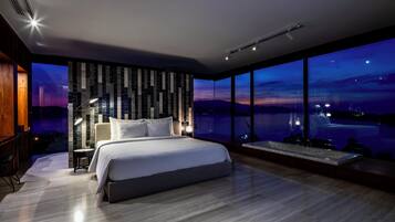 Un dormitorio moderno con una cama grande, una mesita de noche, una lámpara y vistas a un paisaje urbano al anochecer.