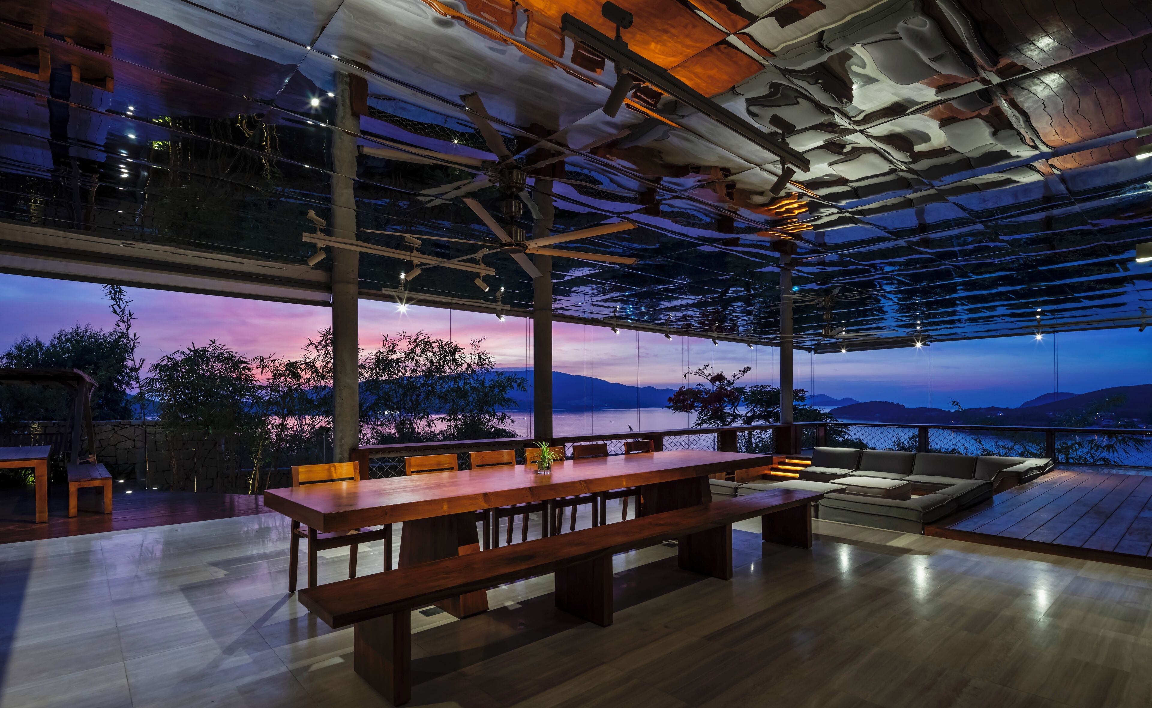 Un vaste espace dédié à la salle à manger, avec une longue table en bois et des bancs, de grandes fenêtres offrant une vue sur un coucher de soleil se déclinant sur les montagnes.