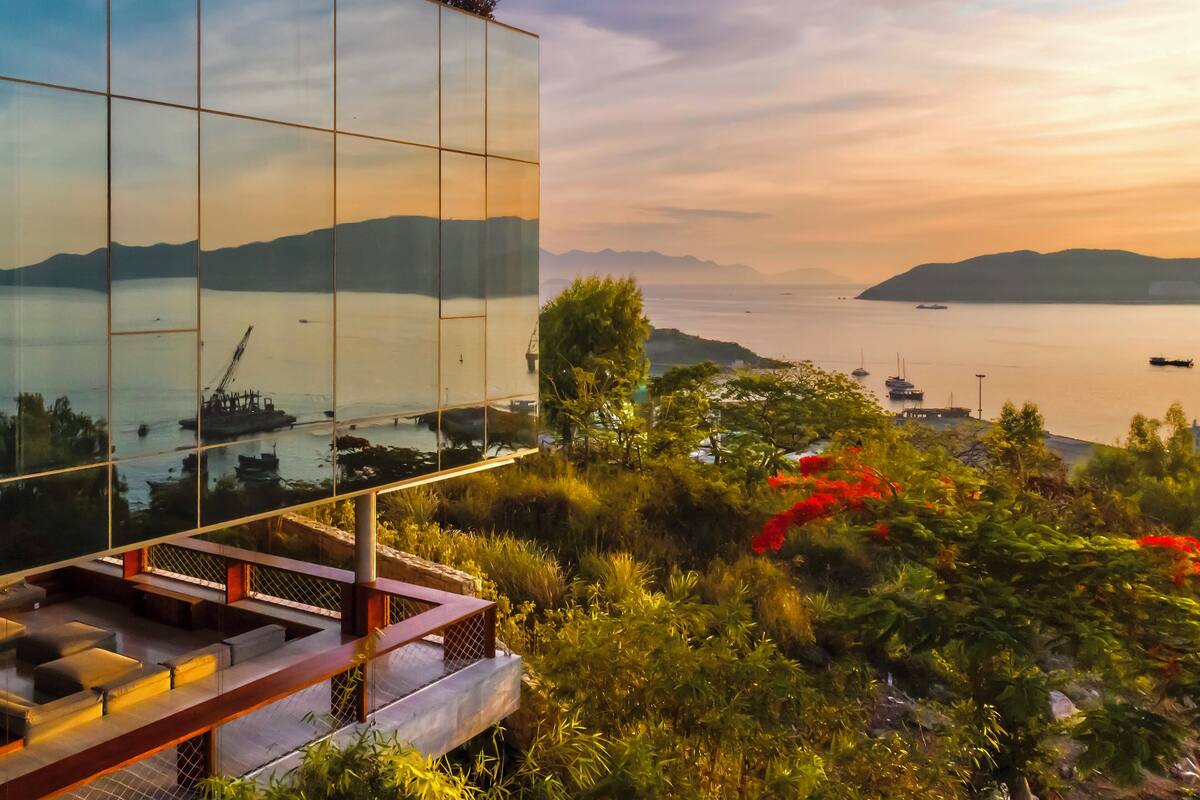 Un bâtiment moderne à la façade de verre qui reflète un coucher de soleil sur une étendue d’eau, entouré d’une végétation luxuriante et de bateaux.