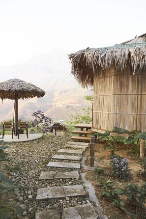 A rustic outdoor seating area with a thatched umbrella, wooden benches, and a stone pathway leading to a bamboo hut. - Eco Hills Homestay (Ta Van)