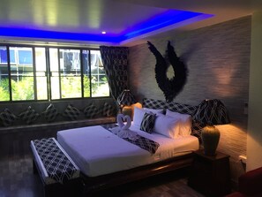 A modern bedroom with a large bed, patterned pillows, a decorative wall piece, and a window with curtains. - Fong Kaew and Baan Nang Fa Guesthouse (Patong)