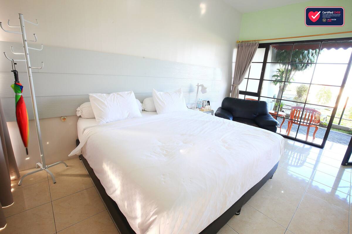A hotel room with a large bed, a chair, a desk with a lamp, and a view of the outdoors through a large window.