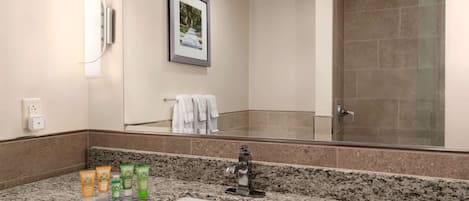 Un baño con encimera de granito, lavamanos, espejo, un cuadro enmarco y zona de ducha.