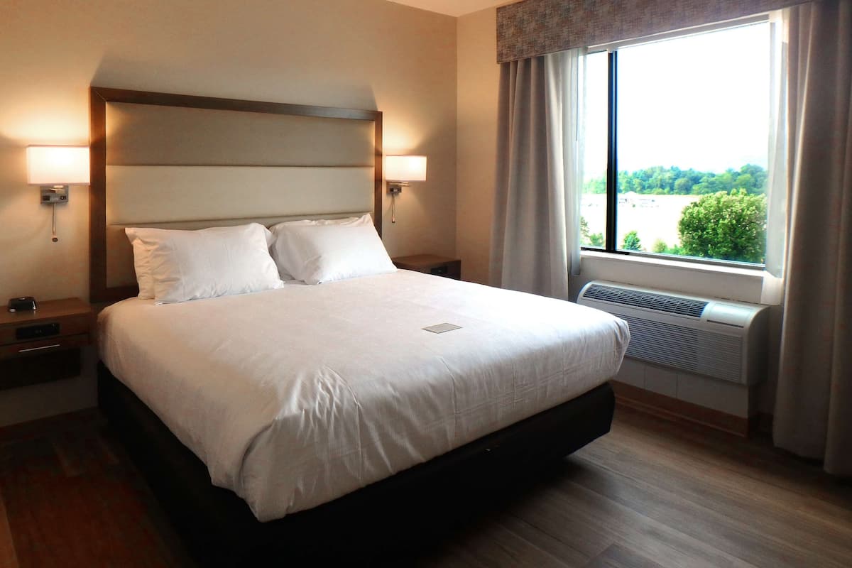 A hotel room with a large bed, two bedside tables, a nightstand, a window with curtains, and an air conditioning unit.