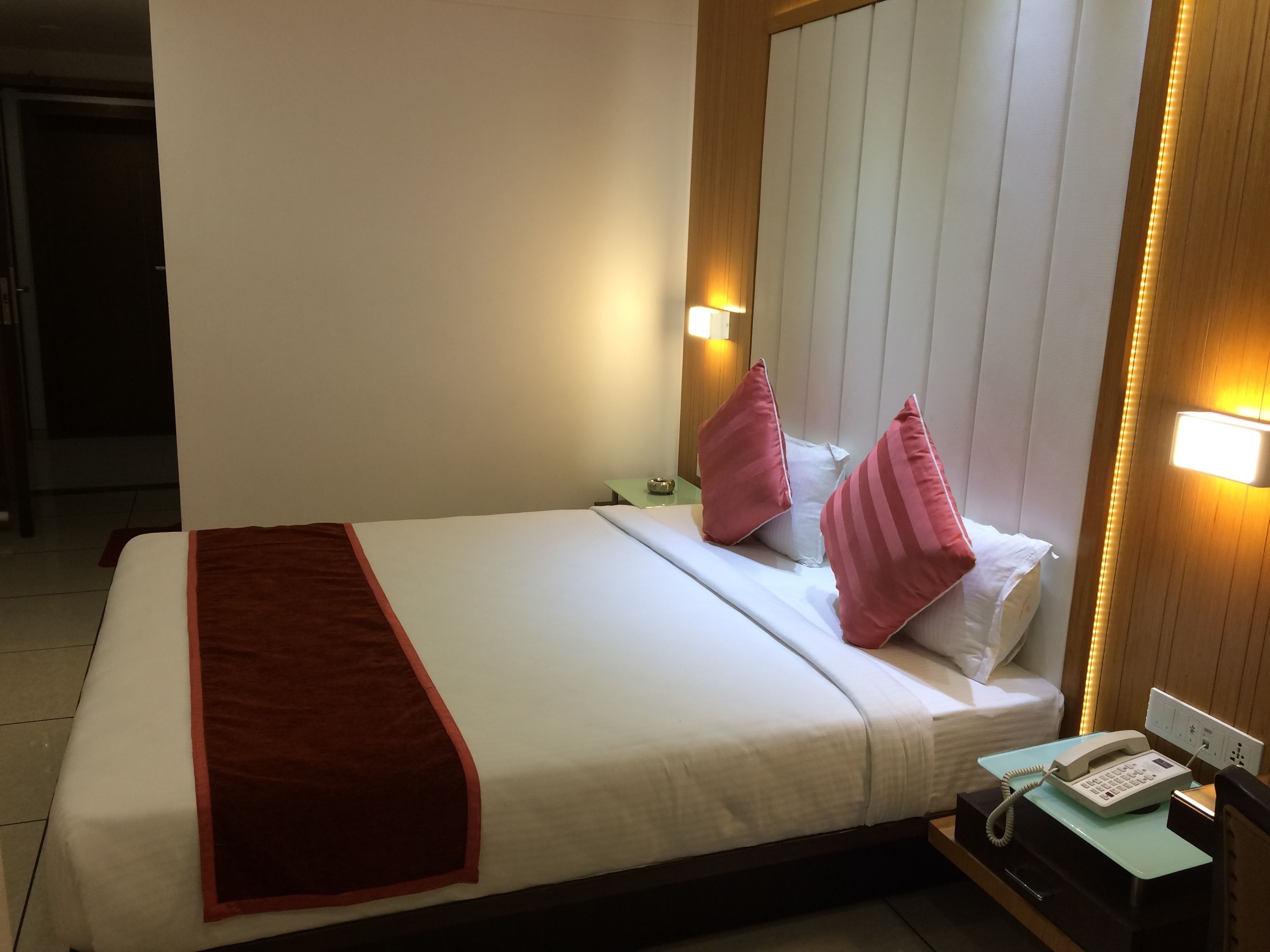 A hotel room with a bed, a bedside table with a phone, and a wall-mounted light fixture.