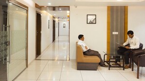 Un hall de hotel con entrada por puerta de vidrio, un área de estar con sofá y sillas, y un pasillo que conduce a otras habitaciones.