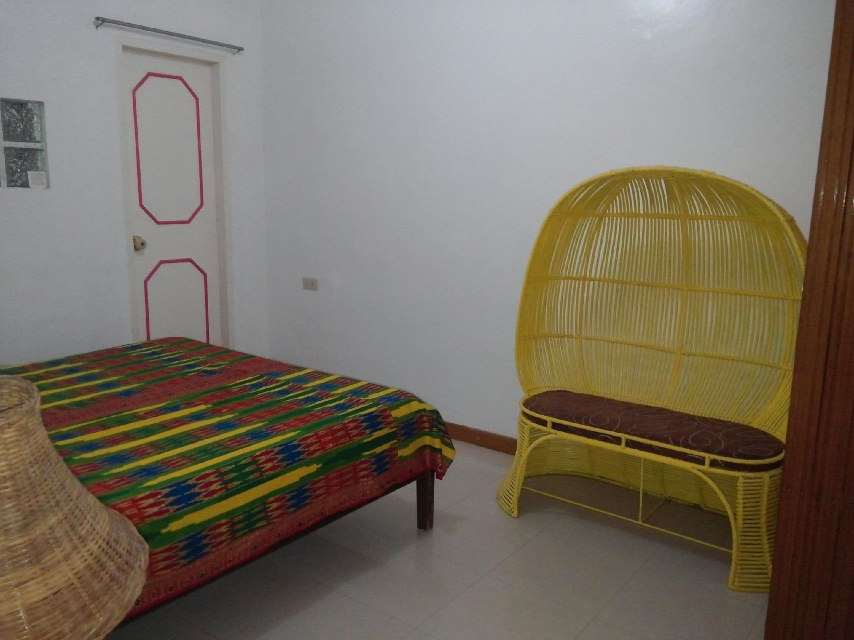 Quarto com uma cama, uma cadeira de vime amarela e uma porta com contorno vermelho.