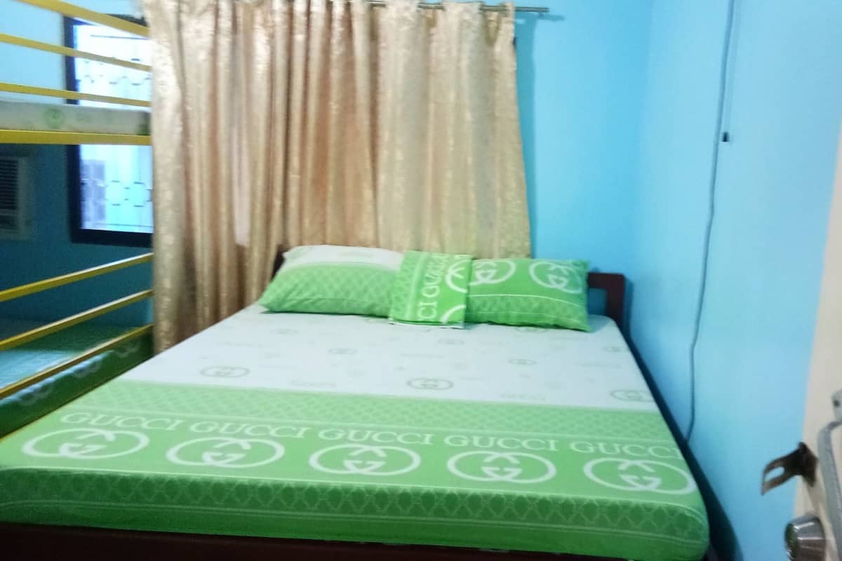 A single bed with a green and white patterned bedspread and pillows, a window with beige curtains, and a blue wall.