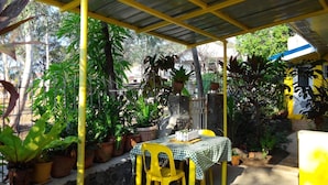 An outdoor patio with yellow chairs and a table covered by a checkered tablecloth, surrounded by potted plants and a metal roof structure. - Yellow HOUSE Vacation Rental (Olongapo)