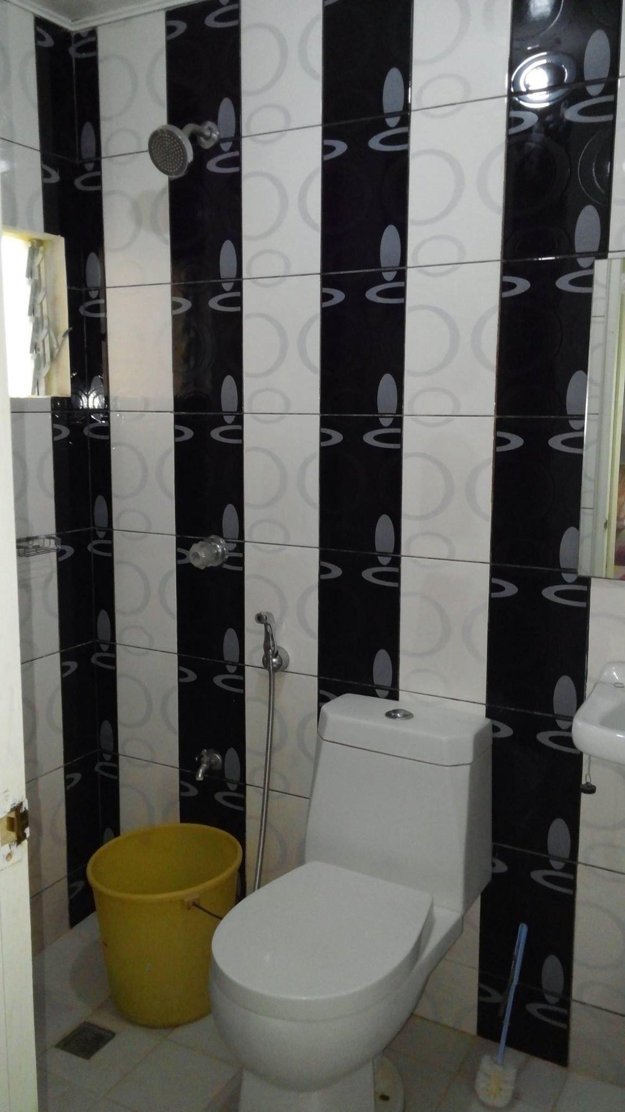 A bathroom with black and white patterned tiles, a white toilet, a yellow bucket, and a handheld showerhead.