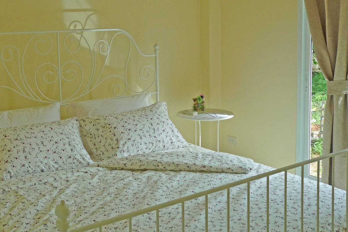 A bed with a white metal frame and floral bedding, a small round table with a vase, and a window with curtains.