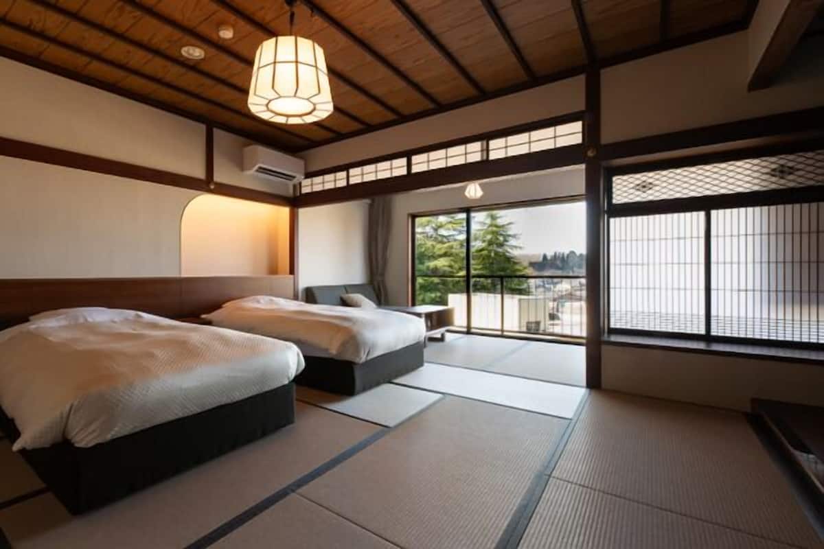 Una habitación de estilo japonés tradicional con suelo de tatami, dos camas, un fusuma que da a un balcón y una lámpara colgante.