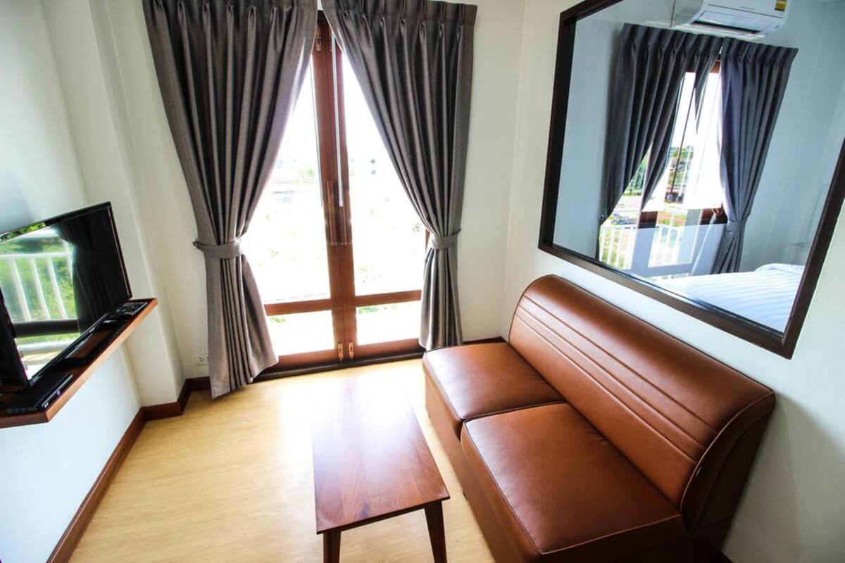 A room with a brown leather sofa, a wooden coffee table, a flat-screen TV on a wooden stand, and a large window with grey curtains.