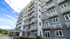 A multi-story residential building with balconies, a parking lot, and a small entrance area with a covered walkway. - Tai-Shan Suites (Ratchaburi)