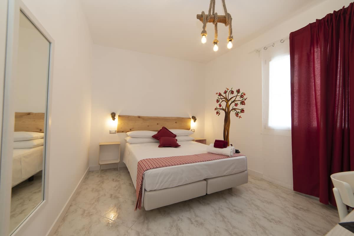 A bedroom with a bed, bedside tables, a mirror, a chandelier, a window with curtains, and a wall-mounted tree decoration.