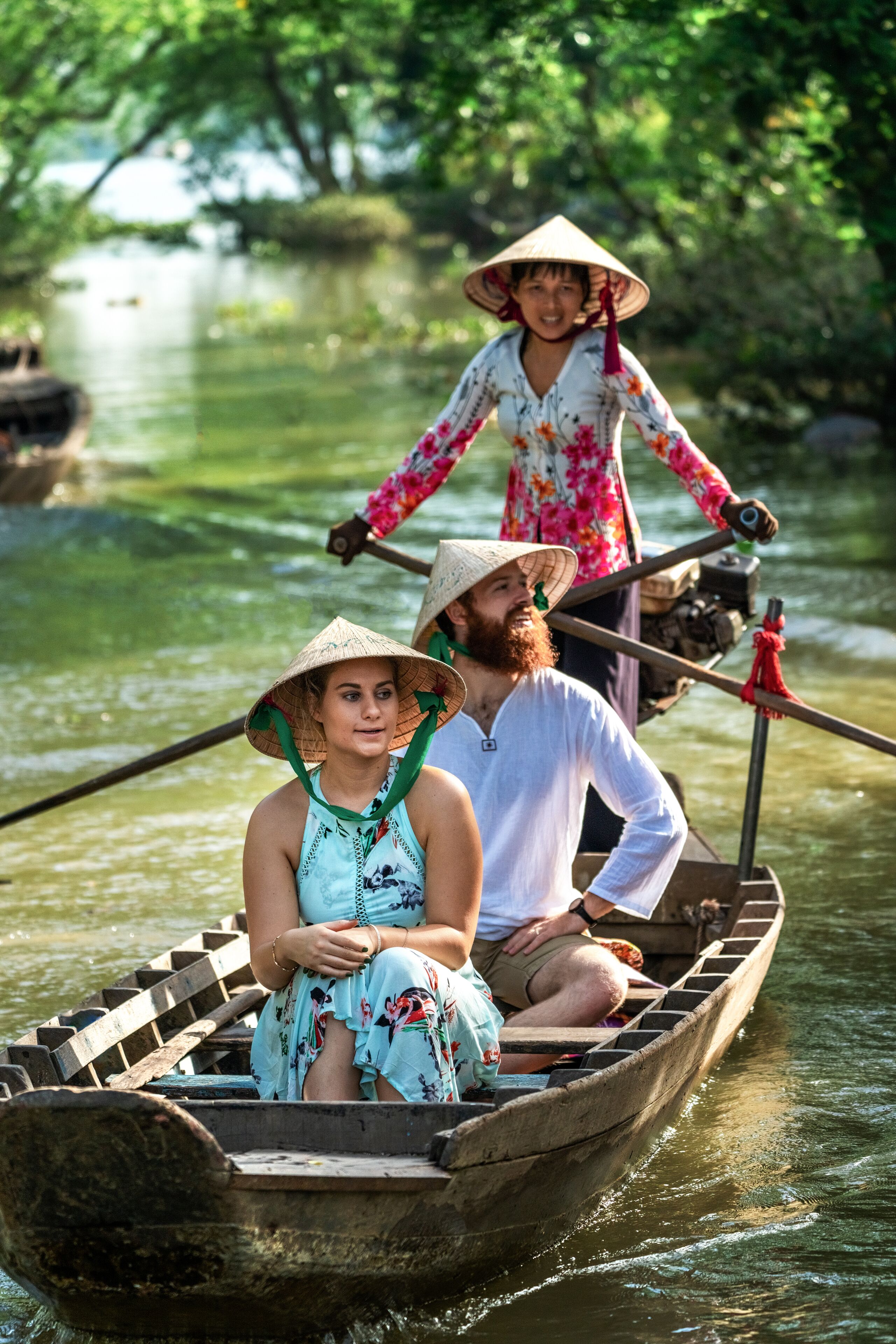 A wooden boat with two people wearing traditional conical hats, one seated and one standing, on a calm river surrounded by greenery.