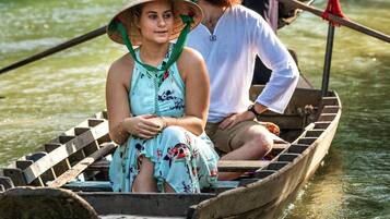 A wooden boat with two people wearing traditional conical hats, one seated and one standing, on a calm river surrounded by greenery.