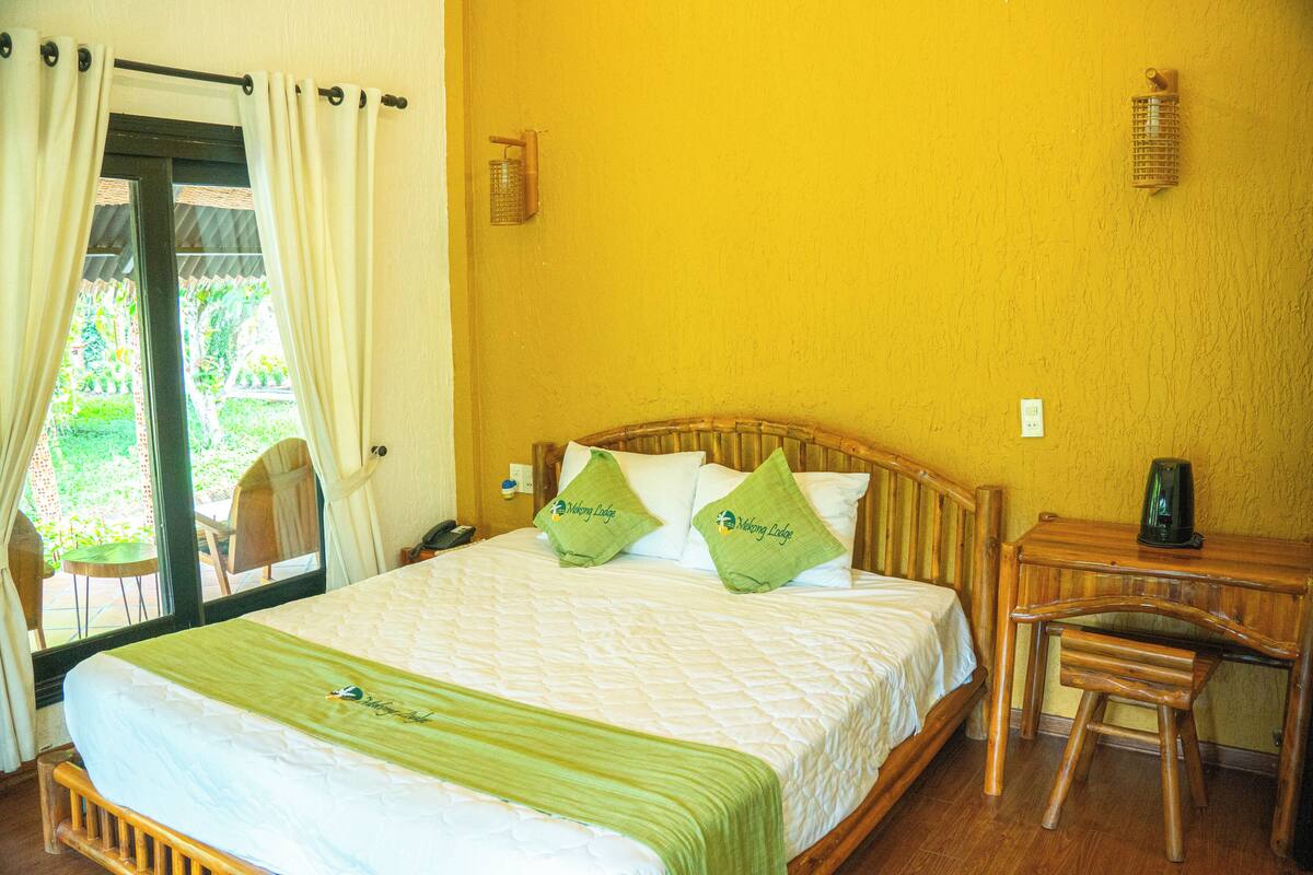 Een bed met een groene spreidek en twee kussens, een houten nachtkastje met een lamp, en een stoel.
