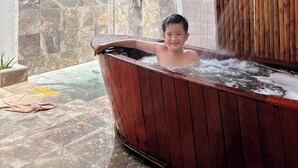 A person in a wooden bathtub with a showerhead, surrounded by stone tiles and a wooden wall. - Mekong Lodge Hotel & Resort (Cai Be)