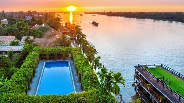 A resort with a swimming pool, lush greenery, and a view of the sunset over the water. - Mekong Lodge Hotel & Resort (Cai Be)