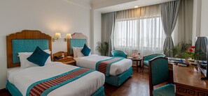 A hotel room with two beds, a desk, a chair, and a view of the city through large windows. - Hotel Le Himalaya by Best Resort Nepal (Kathmandu)