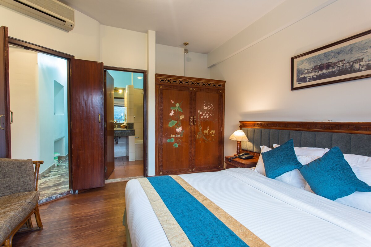 A hotel room with a bed, a wooden wardrobe, a chair, and a bathroom visible through an open door.