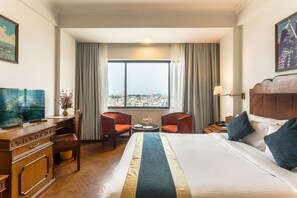A hotel room with a large bed, a desk with a television, two chairs, a small table with flowers, and a view of a cityscape through a large window. - Hotel Le Himalaya by Best Resort Nepal (Kathmandu)