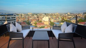 A balcony with two wicker chairs and a small table, overlooking a densely packed cityscape with various buildings and a distant mountain range. - Hotel Le Himalaya (Kathmandu)