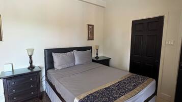 Quarto com cama, mesa de cabeceira, ventilador de teto, porta e quadro na parede.