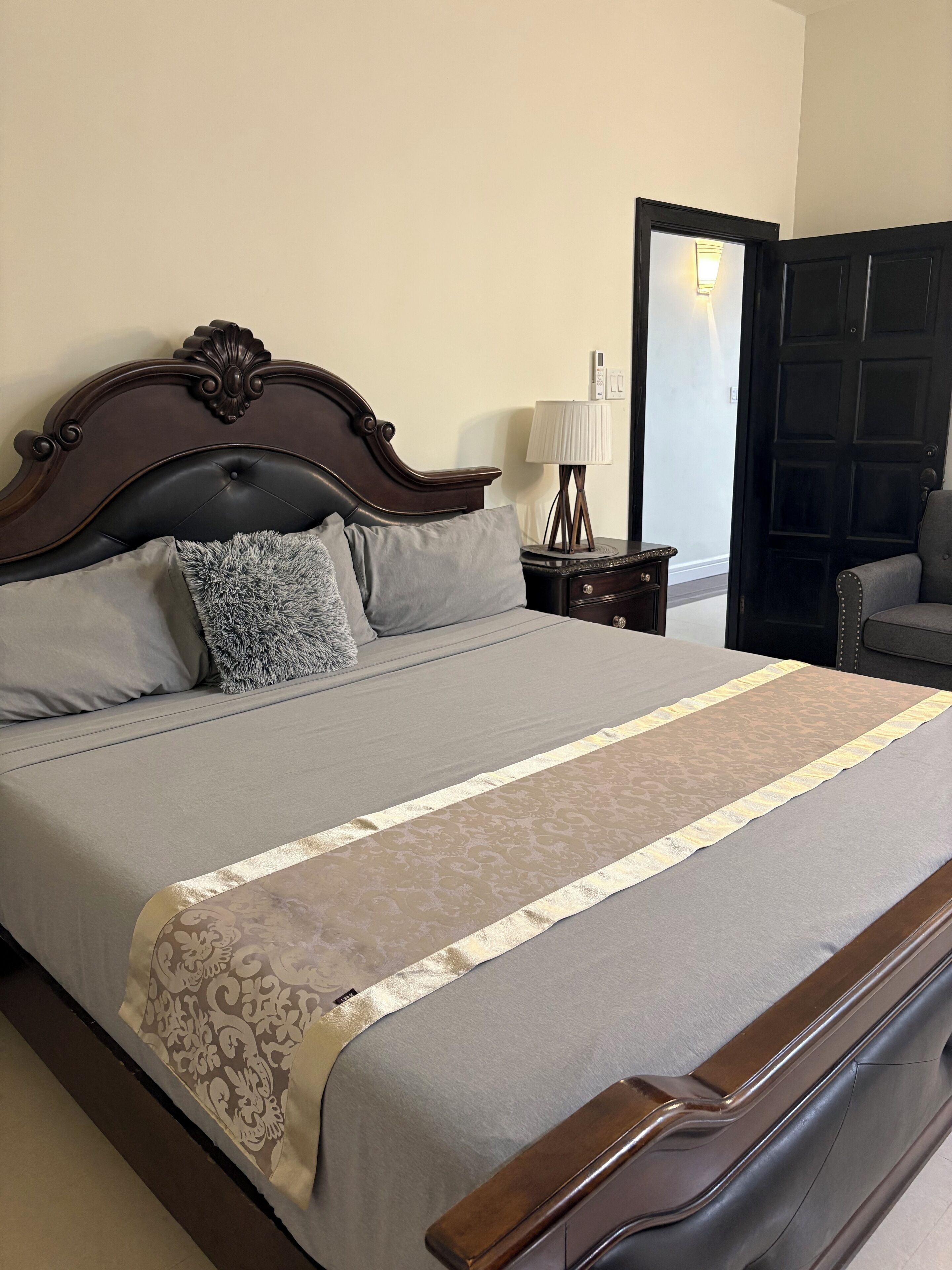 Un lit bien fait, avec une couvre-lit à motifs et des oreillers, une tête de lit en bois, une table de chevet avec une lampe, et une porte visible au fond.
