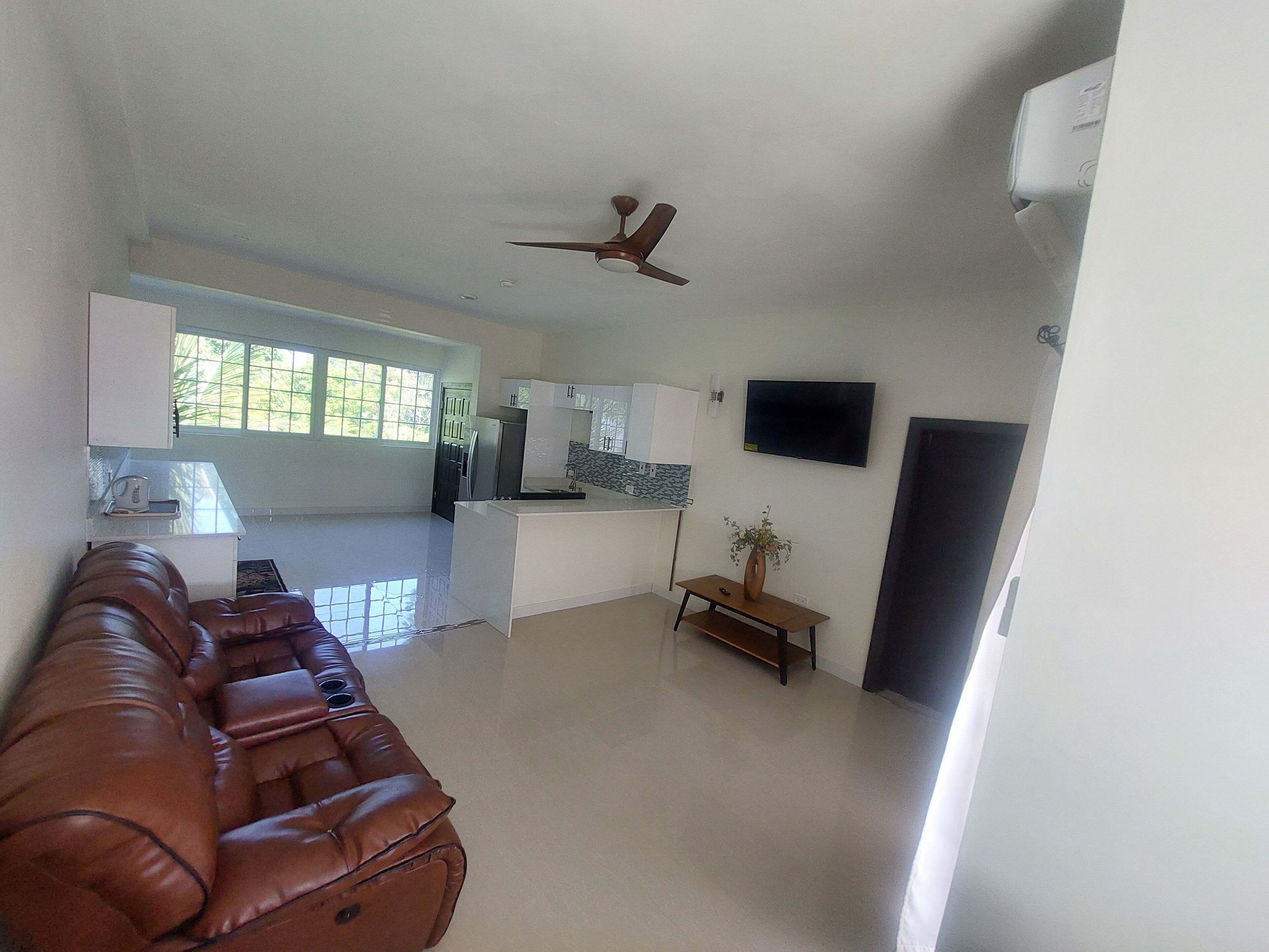 A spacious living area with a brown leather couch, a ceiling fan, a flat-screen TV, and a kitchen area with white cabinets and a window.