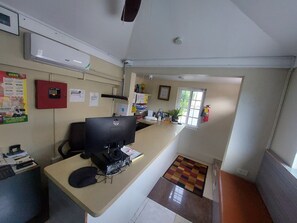 A small office with a desk, computer, and various office supplies. There is a fire extinguisher mounted on the wall and a window with a view of the outside.