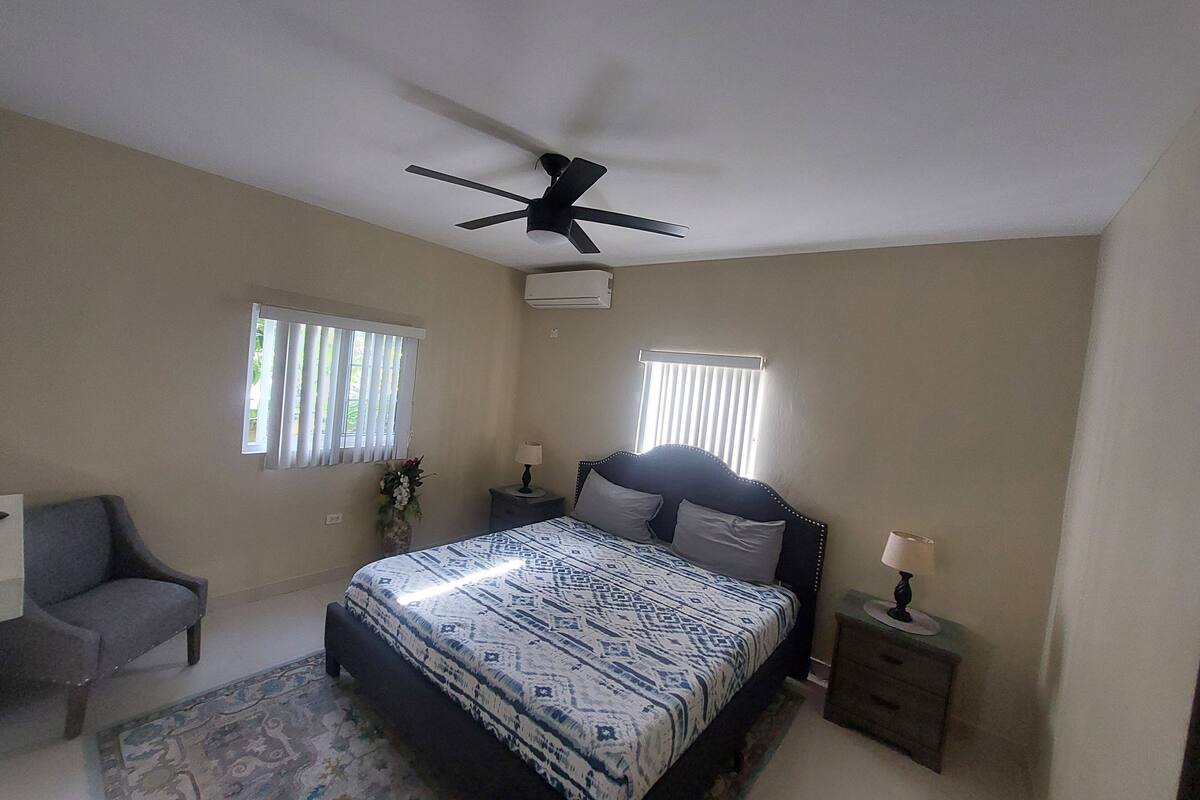 A bedroom with a bed, two bedside tables, a ceiling fan, and two windows with blinds.