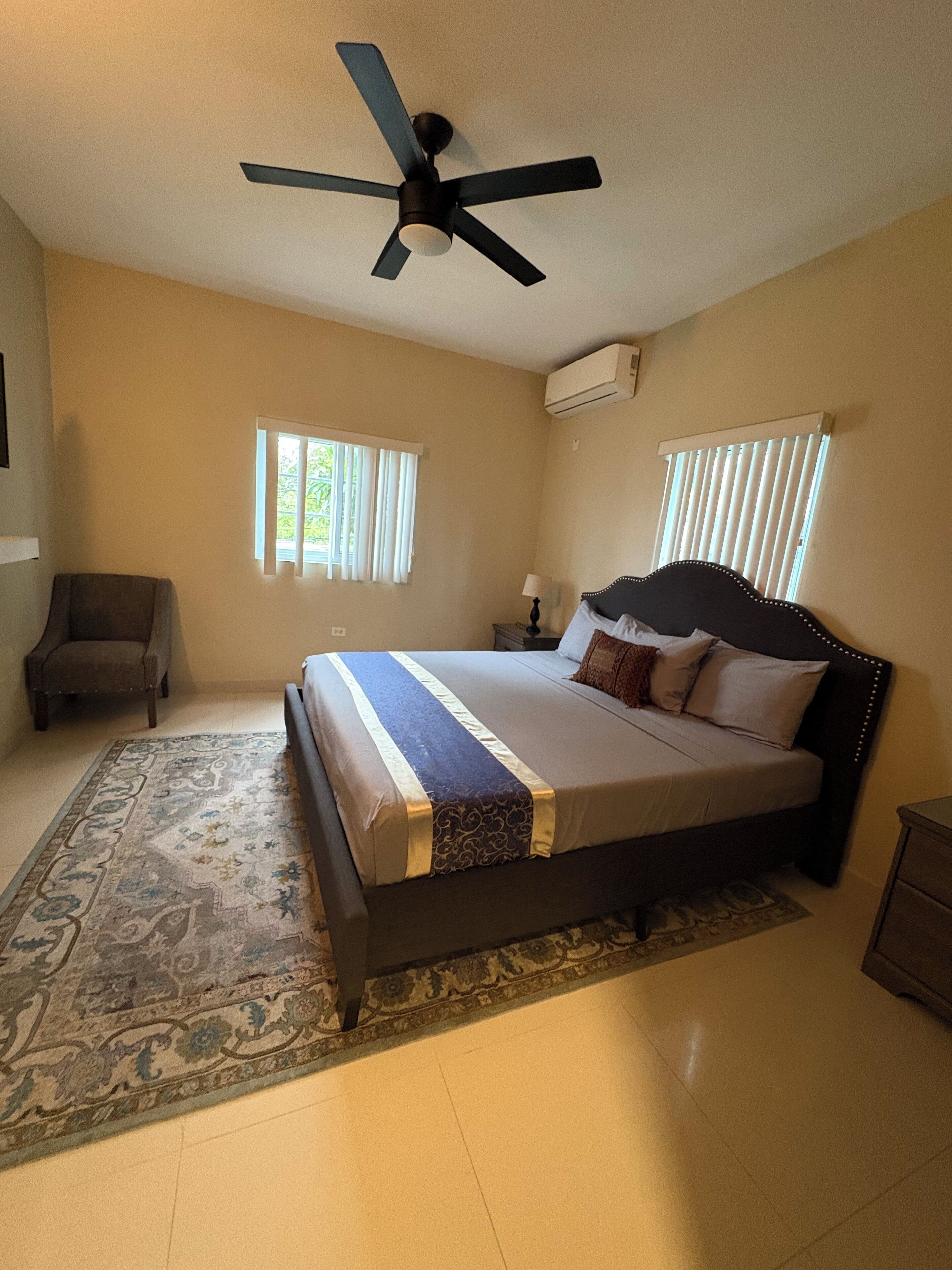 A bedroom with a bed, a ceiling fan, two windows with blinds, a rug, and an air conditioning unit.