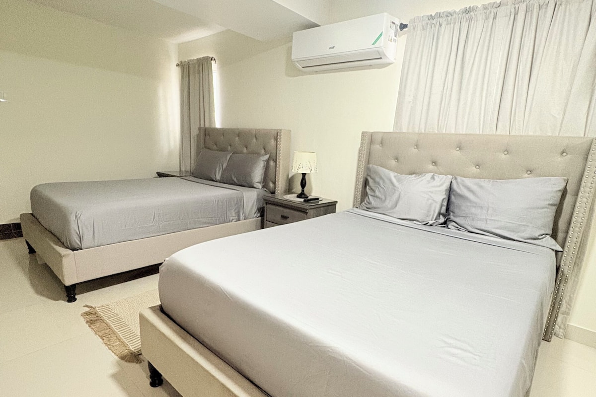 A hotel room with two beds, a nightstand, a lamp, and an air conditioning unit.