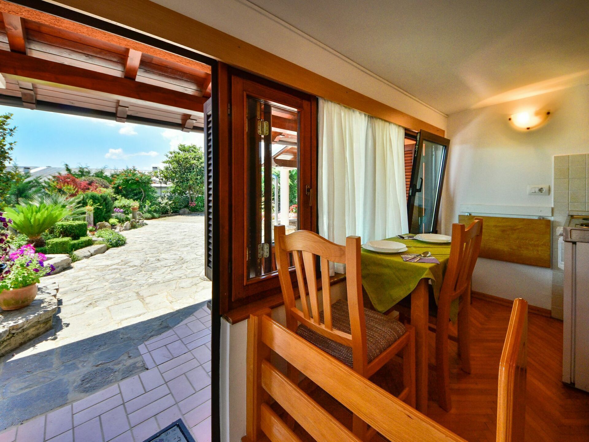 Un coin repas avec une table dressée pour deux personnes, des chaises en bois, une nappe verte et une vue sur un jardin par une porte ouverte.